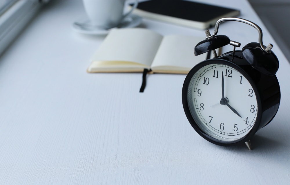 This photo depicts an alarm clock placed on a table with books and a cup of coffee.