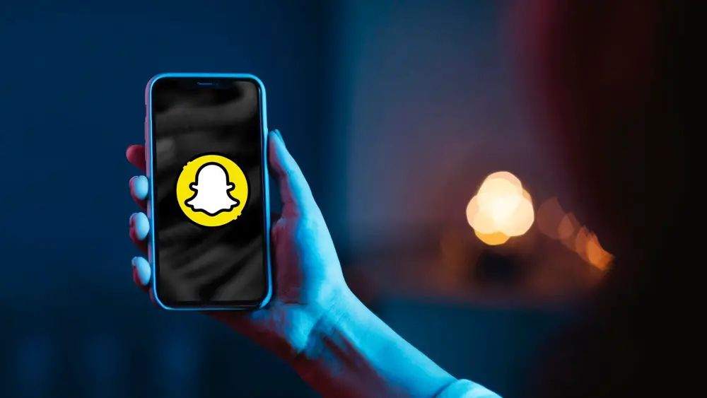 How to Activate Dark Mode on Snapchat [iOS and Android]: This photo depicts a hand holding a smartphone with the Snapchat icon on its screen against a dark background.