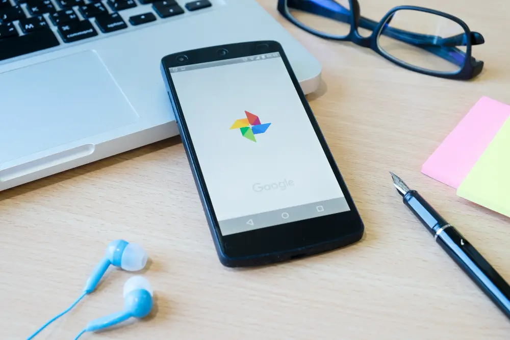 How to Stop Auto Backup on Google Photos: This photo shows a smartphone with its screen featuring the Google Photos logo. The phone is placed on an open laptop, with a pair of reading glasses, a pen, and an earphone nearby.