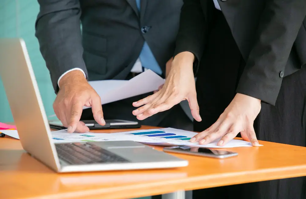 This photo depicts 2 businessmen looking at some business statistics, with a smartphone, a tablet, and a laptop on the desk.