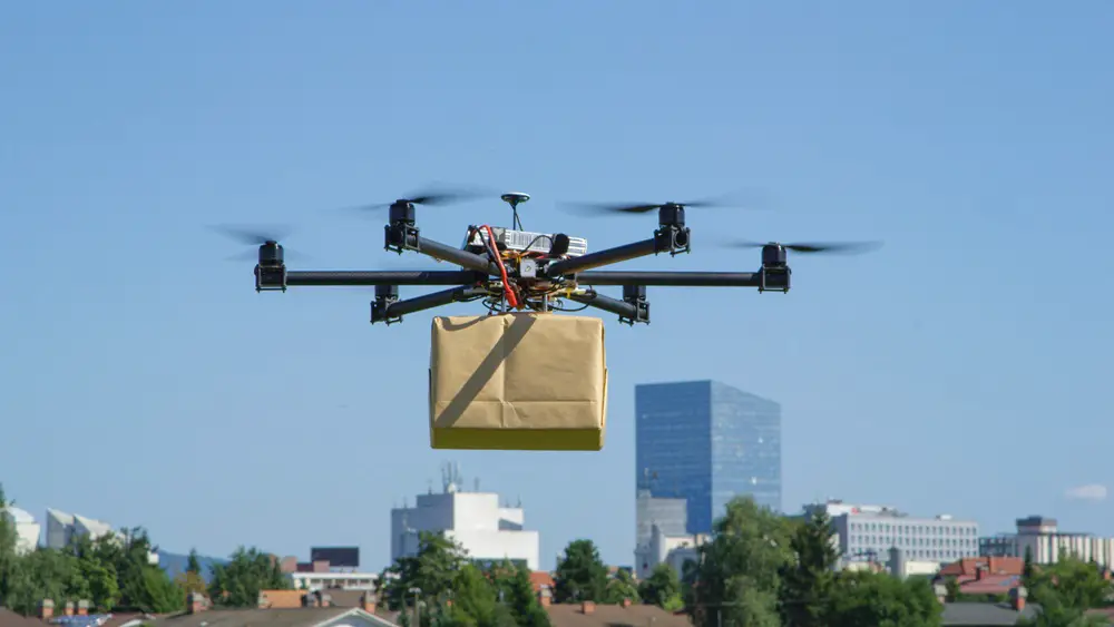 This photo depicts a drone delivering a brown package to an urban city.