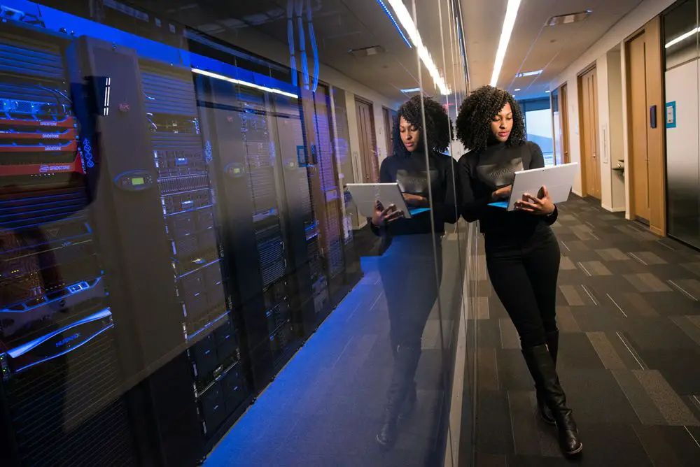This photo depicts a woman standing outside a room with glass walls, where servers are neatly arranged on server racks. The glass walls allow visibility into the interior from the outside.