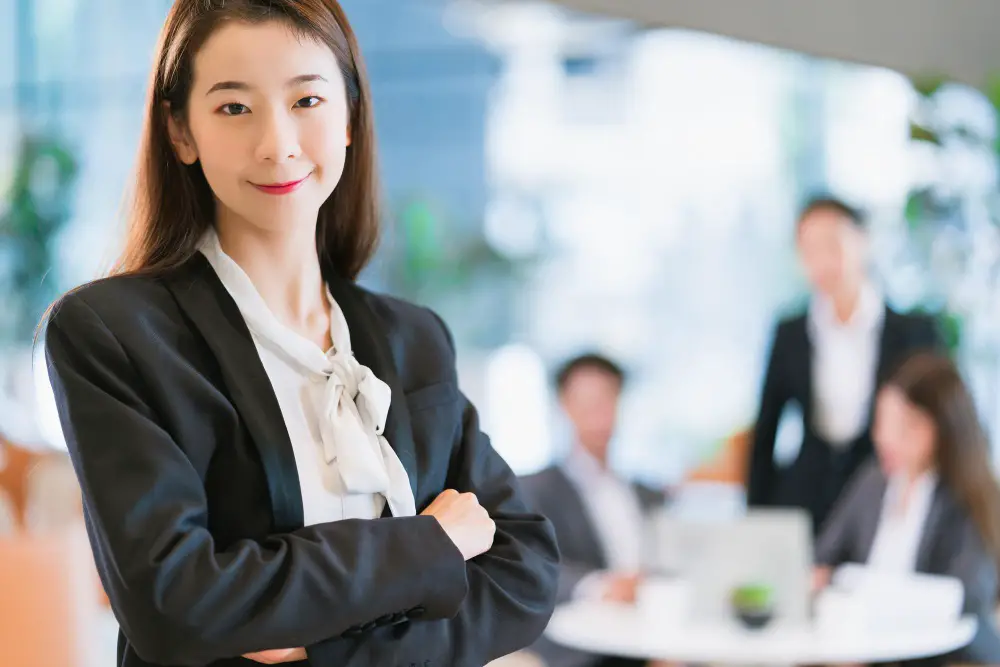 This photo depicts an Asian businesswoman smiling with confidence and leadership in a modern office background.
